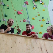 Kletterzentrum Drei junge Erwachsene lächeln und zeigen Daumen hoch vor einer bunten Kletterwand.