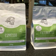 Neanderthaler Kaffee aus der Kult-Kaffee Rösterei in Heiligenhaus Zwei Kaffeebeutel aus der Kult-Kaffee Rösterei, links Espresso, rechts Filterkaffee, vor Regal.