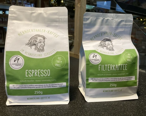 Neanderthaler Kaffee aus der Kult-Kaffee Rösterei in Heiligenhaus Zwei Kaffeebeutel aus der Kult-Kaffee Rösterei, links Espresso, rechts Filterkaffee, vor Regal.