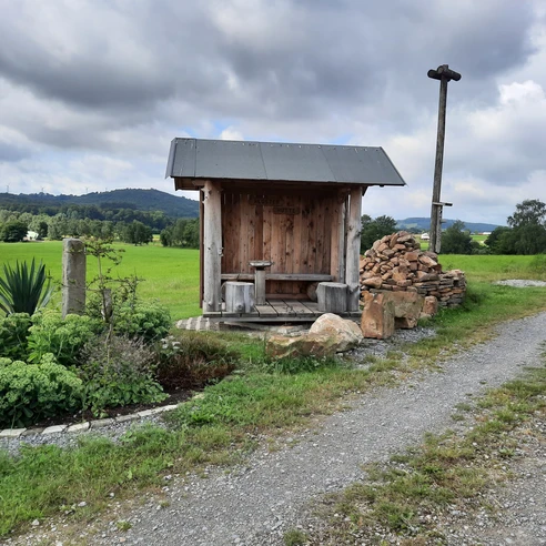 Eine rustikale Holzhütte mit Sitzbank am Wegesrand, umgeben von grünen Feldern und Hügeln.