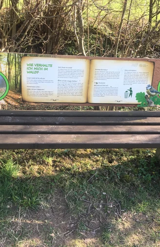 Eine Parkbank mit Infotafel zum verantwortungsvollen Verhalten im Wald, umgeben von Büschen.
