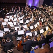 Bläserphilharmonie Aachen, bestehend aus vielen Musiker*innen in eleganter Abendgarderobe, die konzentriert ihre Instrumente spielen. Die Mitglieder des Orchesters sitzen in Formationen, die typische Orchesterinstrumente wie Trompeten, Hörner und Klarinetten umfassen. Der Dirigent steht vorne.