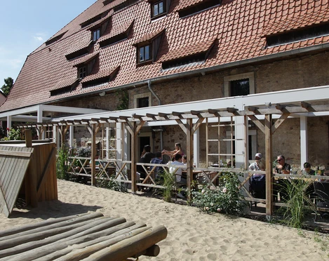 Außenbereich vom Alten Brauhaus zu Fallersleben Der Außenbereich des Alten Brauhauses zu Fallersleben zeigt einen gemütlichen Biergarten mit Holztischen unter einer Pergola. Links ist ein kleiner Holzspielplatz auf sandigem Boden zu sehen. Der Backsteinbau im Hintergrund trägt ein rotes Ziegeldach mit Gauben.
