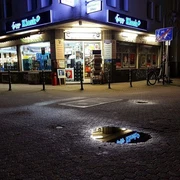 Jazz Kiosk Der Jazz-Kiosk bei Nacht zeigt ein beleuchtetes Geschäft in urbaner Umgebung mit reflektierendem Schaufenster und Regenpfütze.The jazz kiosk at night shows an illuminated store in an urban environment with a reflective shop window and a puddle of rain.