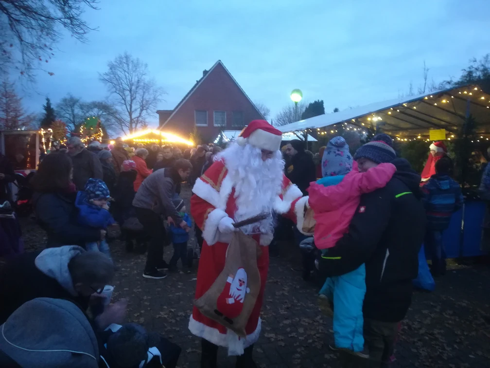 Weihnachtsmarkt in Apen.jpg Weihnachtsmann verteilt Geschenke an Kinder auf belebtem Markt mit Lichtern und Buden in Apen.