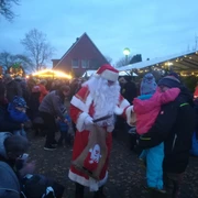 Weihnachtsmarkt in Apen.jpg Weihnachtsmann verteilt Geschenke an Kinder auf belebtem Markt mit Lichtern und Buden in Apen.