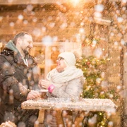 markt-im-advent-schneehuette.jpg Eine Frau und ein Mann lachen an einem Holztisch im Schnee, umgeben von warmem Licht und Winterdekorationen.