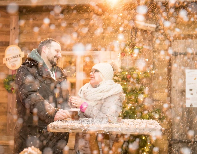 markt-im-advent-schneehuette.jpg Eine Frau und ein Mann lachen an einem Holztisch im Schnee, umgeben von warmem Licht und Winterdekorationen.