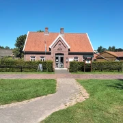 IMG20220811124054.jpg Das Bild zeigt ein charmantes Backsteinhaus mit rotem Ziegeldach inmitten einer grünen Wiese unter einem strahlend blauen Himmel. Vor dem Haus sind gepflegte Büsche und ein gepflasterter Weg sichtbar, der zur Eingangstür führt.
