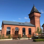 Außenbereich Eisenhütte Das Bild zeigt ein historisches, rotes Backsteingebäude. Es hat große Fenster und einen markanten, spitzen Turm. Der Himmel ist klar und blau.