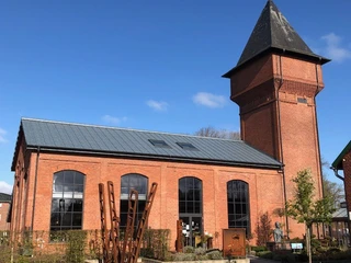 Außenbereich Eisenhütte Das Bild zeigt ein historisches, rotes Backsteingebäude. Es hat große Fenster und einen markanten, spitzen Turm. Der Himmel ist klar und blau.