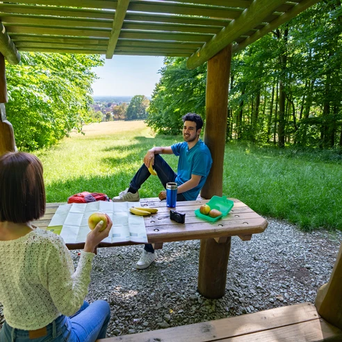 Zwei Personen sitzen an einem Holztisch unter einem hölzernen Pavillon im sommerlichen Wald.