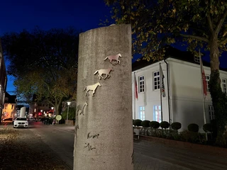 Hufeisenspur Verden Relief von Pferden auf einer Steinsäule bei Nacht, flankiert von Bäumen und beleuchteten Gebäuden.