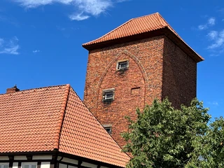 Alter Wehrturm Verden Backsteinturm mit roten Ziegeln, darüber ein blauer Himmel; teilverdeckte Fachwerkhäuser im Vordergrund.