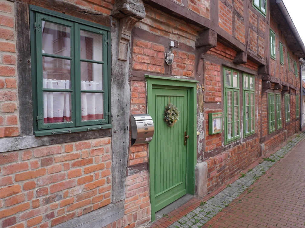 Fachwerkhäuser mit roten Ziegelwänden, weißen Fenstern und grünen Türen entlang einer Pflastergasse.