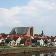 Eine mittelalterliche Kathedrale überragt ein Dorf mit roten Dächern am Ufer eines ruhigen Flusses.