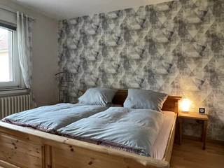ferienwohnung_hermanns_schlafzimmer.jpg Schlafzimmer mit Doppelbett aus Holz, blauer Bettwäsche, gemusterter Tapete und kleinem Nachttisch.
