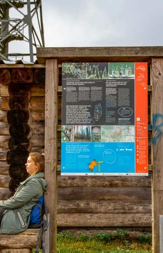 Infotafel am Baumweg Frau sitzt entspannt vor einem hölzernen Wegweiser mit Informationstafel, im Hintergrund ein Turm.