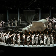 Cologne Opera in the StaatenHaus Eine Bühne voller grau-weißer Kostüme, ein Chor dahinter; eine monumentale Kopfskulptur dominiert.A stage full of gray and white costumes, a choir behind it; a monumental head sculpture dominates.