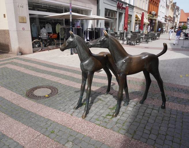 Skulptur Fohlengruppe Verden Bronzeskulptur zweier Fohlen in einer belebten Fußgängerzone von Verden mit Geschäften im Hintergrund.