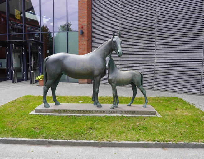Skulptur Mutterstute mit Fohlen Verden Bronzeskulptur einer Pferdestute mit ihrem stehenden Fohlen auf einem Sockel vor einem modernen Gebäude.