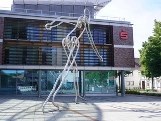 Walking Man Verden Die Skulptur "Walking Man" aus Metallstäben steht vor einer modernen Bank mit großen Glasfenstern.