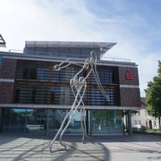 Walking Man Verden Die Skulptur Walking Man aus Metall steht vor einem modernen Gebäude mit Glasfassade in Verden.