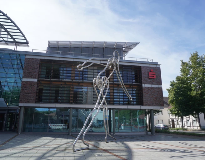 Walking Man Verden Die Skulptur Walking Man aus Metall steht vor einem modernen Gebäude mit Glasfassade in Verden.