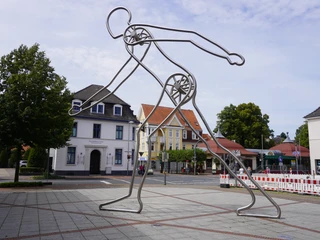 Skulptur Walking Man Verden Moderne Metallskulptur eines gehenden Mannes in Verden, umgeben von Gebäuden und Bäumen.