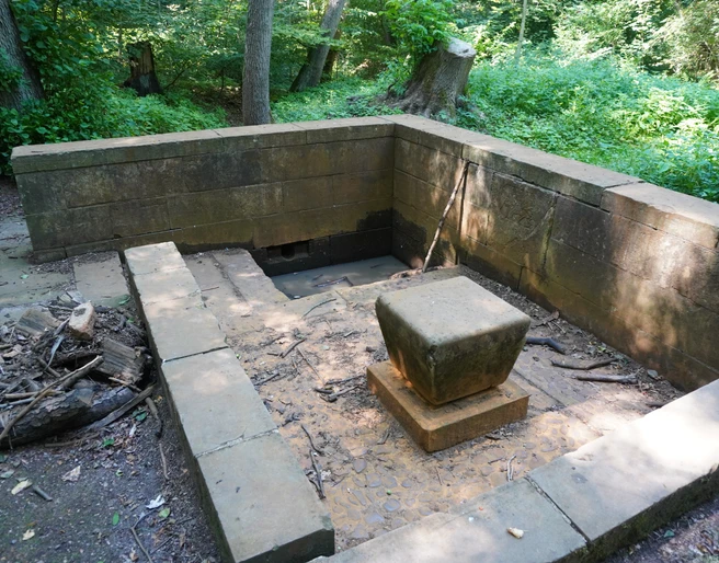 Ein steinerner Brunnen im Wald mit quadratischem Sockel, umgeben von Bäumen und dichtem Grün.