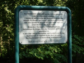 Eine grüne Infotafel im dichten Wald beschreibt die Geschichte des Verdener Brunnens ausführlich.
