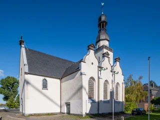 Saint Clemens Die Sankt Clemens Kirche ist ein weißes, historisches Gebäude mit markantem Turm und schmuckvollen Fenstern. Links sind Bäume und blauer Himmel zu sehen.St. Clement's Church is a white, historic building with a striking tower and ornate windows. Trees and blue sky can be seen on the left.
