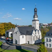 Saint Clemens Das Bild zeigt die Sankt Clemens Kirche in einer luftigen Perspektive, eingebettet in eine grüne städtische Umgebung. Die weiße Kirche mit schwarzem Dach und charakteristischem Turm steht neben einem historischen Gebäude bei sonnigem Wetter.The picture shows St. Clement's Church from an airy perspective, embedded in a green urban environment. The white church with its black roof and characteristic tower stands next to a historic building in sunny weather.