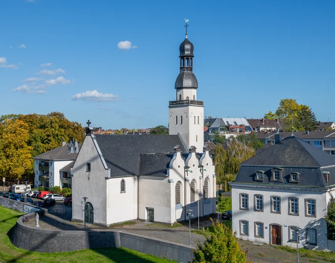 Saint Clemens Das Bild zeigt die Sankt Clemens Kirche in einer luftigen Perspektive, eingebettet in eine grüne städtische Umgebung. Die weiße Kirche mit schwarzem Dach und charakteristischem Turm steht neben einem historischen Gebäude bei sonnigem Wetter.The picture shows St. Clement's Church from an airy perspective, embedded in a green urban environment. The white church with its black roof and characteristic tower stands next to a historic building in sunny weather.