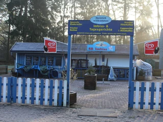 Magic Park Verden Ein blau-weiß gestrichenes Imbisshäuschen im Freien, umgeben von Bäumen und Sandweg, lädt mit kindgerechten Speisen und Getränken zum Verweilen ein.