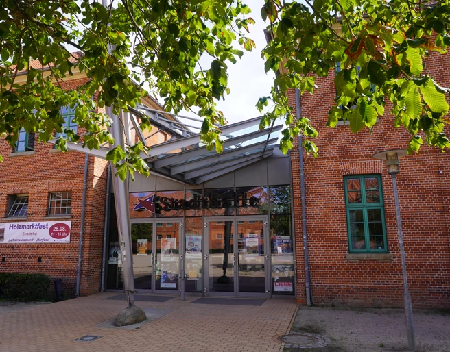 Eingang der Stadthalle Verden mit Glasfassade und rotem Ziegelbau, umrahmt von Kastanienblättern.