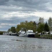 Motorboote liegen am ruhigen Flussufer, umgeben von Bäumen unter einem bewölkten Himmel.