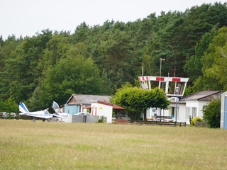 Ein Sportflugzeug parkt auf einem grasbewachsenen Flugplatz vor einem kleinen weißen Flughafengebäude.
