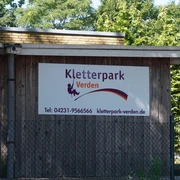 Holzgebäude mit Schild des Kletterparks Verden, umgeben von Bäumen, bietet Abenteuer und Natur.