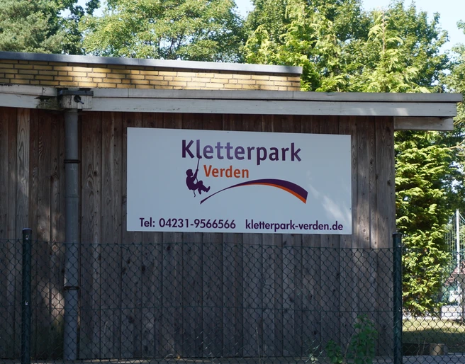 Kletterpark Verden Holzgebäude mit Schild des Kletterparks Verden, umgeben von Bäumen, bietet Abenteuer und Natur.