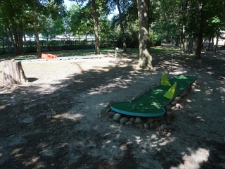 Minigolfbahn im Schatten hoher Bäume in einem Waldgebiet, mit gelben Hindernissen und natürlichem Umfeld.