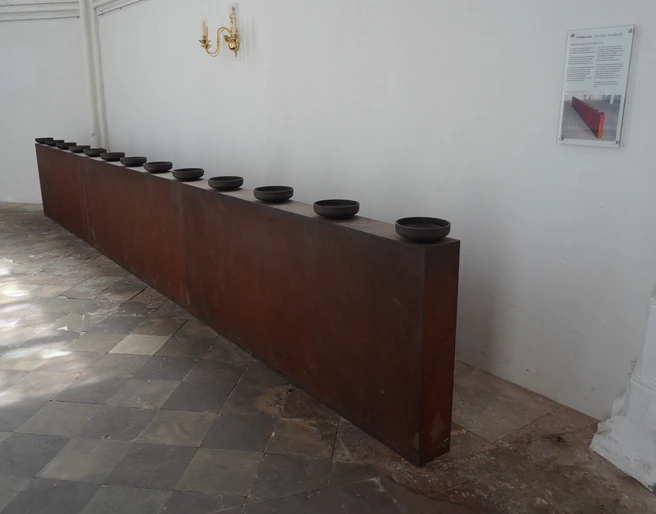 Skulptur Das letzte Abendmahl Verden Die Skulptur zeigt zehn dunkle Schalen auf einem langen, rechteckigen Metallblock in einem Innenraum.