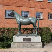 Tempelhüter Bronzestatue eines Pferdes namens Tempelhüter vor roter Backsteinfassade, umgeben von Büschen.