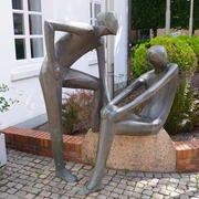 Skulptur vor VGH Moderne Bronzeplastik zweier stilisierter Figuren auf einem Granitstein, umrahmt von Pflanzen und Büschen.
