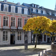 Elegantes Backsteingebäude mit hohen Fenstern und einem gepflegten Vorplatz, wird von einem goldgelben Baum und einem Brunnen vom Hühnerdieb geschmückt. Im Hintergrund erhebt sich der Turm einer Kirche.