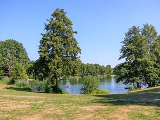 Grüner Park mit See, umgeben von Bäumen, zwei Personen paddeln auf dem Wasser.