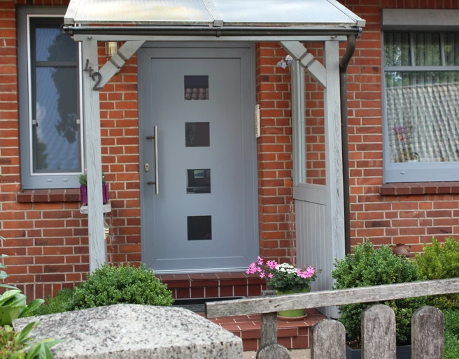 FeWo Wehrmann Eingang einer Ferienwohnung mit verglastem Vordach, modernen grauen Tür, Blumendekor und Ziegelbau.