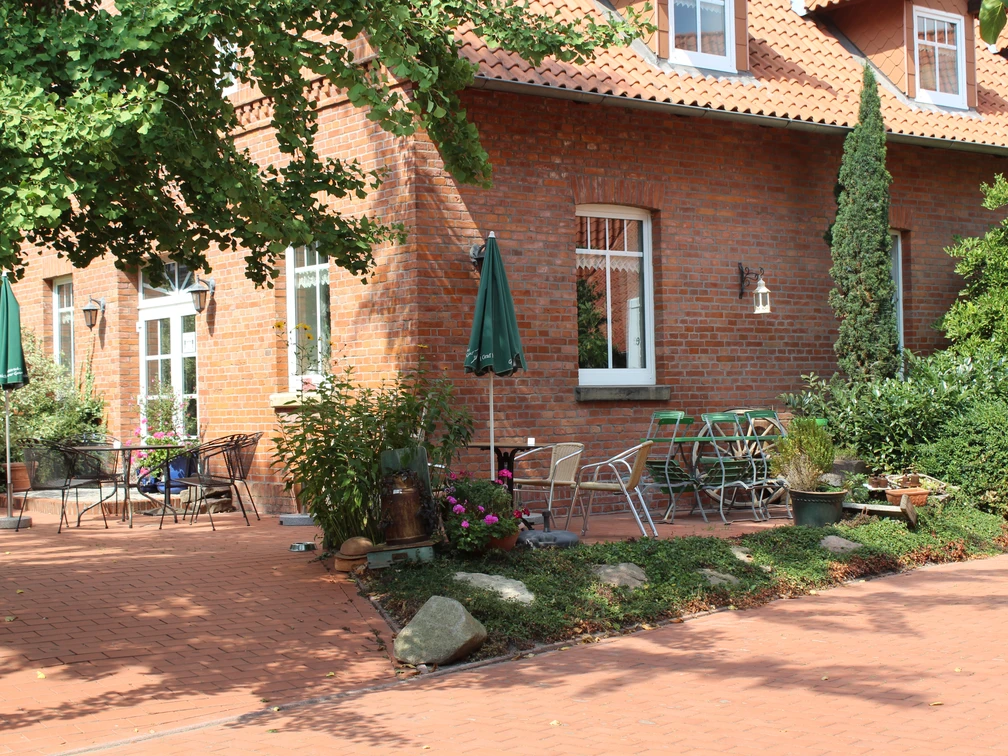 Kleine Kaffeediele Roter Klinkerbau mit Terrasse, Gartenmöbeln und grünen Pflanzen unter blauen Sonnenschirmen.