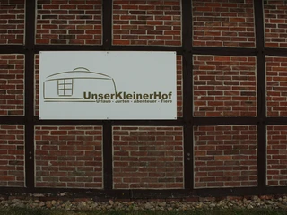 Unser kleiner Hof  Ein Schild an einer Backsteinwand mit der Aufschrift "Unser Kleiner Hof - Urlaub - Jurten - Abenteuer - Tiere".