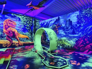 Blacklight_District2_foto_ccbysa_foto©adventure-district.jpeg Das Bild zeigt einen Dinosaurier-Themen-Minigolfplatz im Neonlicht, umgeben von Wandgemälden prähistorischer Tiere und Pflanzen.